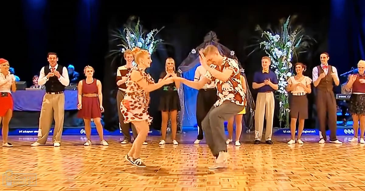 A dance performance in what appears to be a ballroom setting, with two dancers prominently in the foreground: one wearing a leopard print dress and black sneakers, another in a Mickey Mouse shirt with light-colored shoes. Other individuals stand in a line in the background clapping or observing.