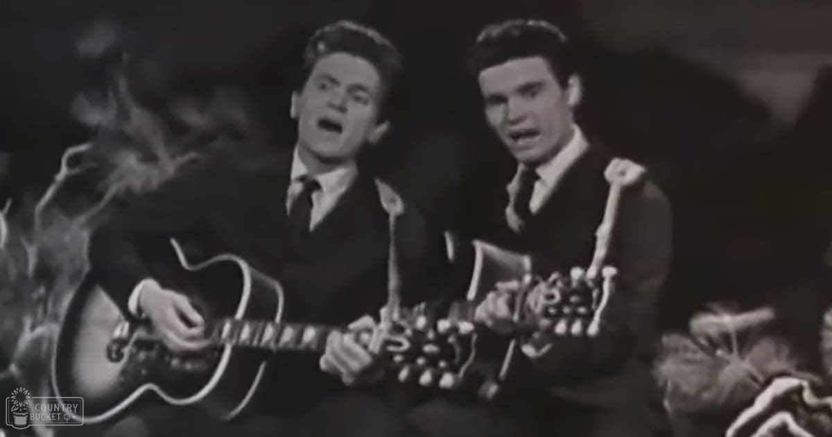 Two young adult males performing music in a dimly lit studio setting with dark tones.