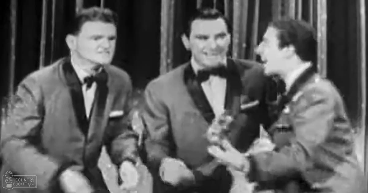 Three adult males in formal suits, possibly from a mid-20th-century entertainment scene, indoors with curtains and monochromatic lighting.