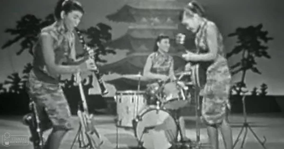A musical performance scene on a stage set against an outdoor backdrop, likely summer in a tropical or Asian theme with performers wearing sleeveless dresses and styled hair.