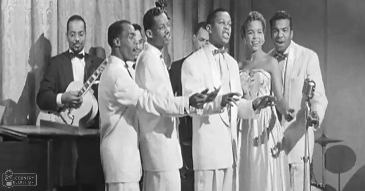 A mid-20th century musical performance group indoors with performers wearing suits and holding instruments, piano and drum set partially visible.
