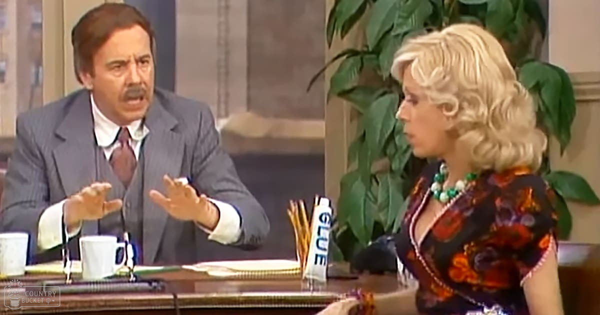 A middle-aged man wearing a dark gray suit and maroon tie interacts with a woman in a floral-patterned blouse, both seated at an office desk filled with various items like mugs, papers, glue, and a microphone.