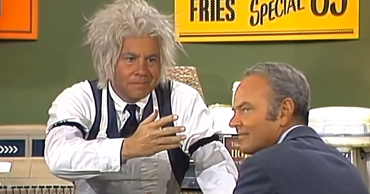 Two middle-aged men in a diner setting, one wearing a white shirt and dark vest, the other in a dark blue suit, with signs reading 'FRIES SPECIAL' overhead.