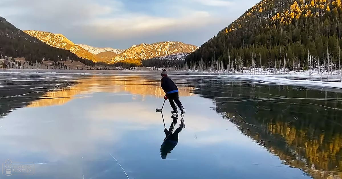 An adult male wearing a dark jacket with light blue accents, black pants, ice skates, and a white knit hat is ice skating on a frozen lake surrounded by snow-covered mountains in winter.