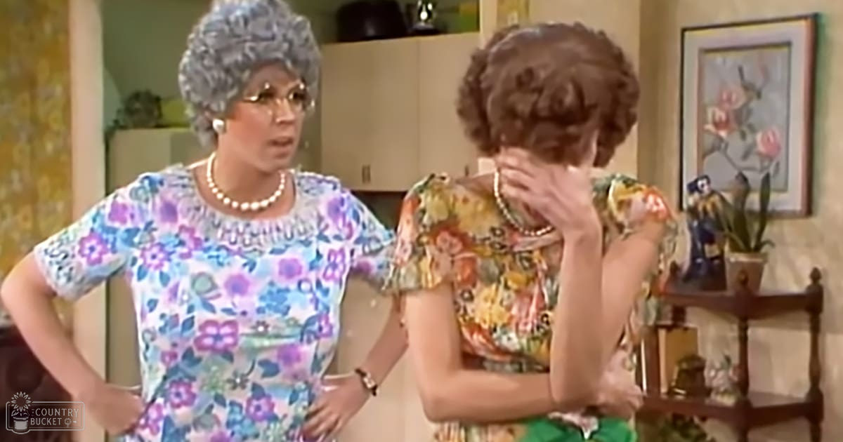 Two older adult females wearing floral dresses in a warm, indoor setting with light-colored wallpaper and wooden furniture.