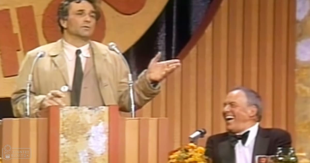 A man in a beige blazer and white shirt speaking at an event, with another older adult seated beside him wearing a dark suit jacket over a light dress shirt.