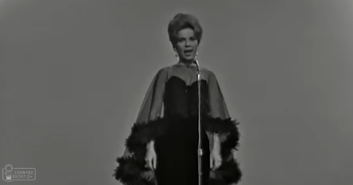 Adult female in a black dress with feathered cape in an indoor studio setting.