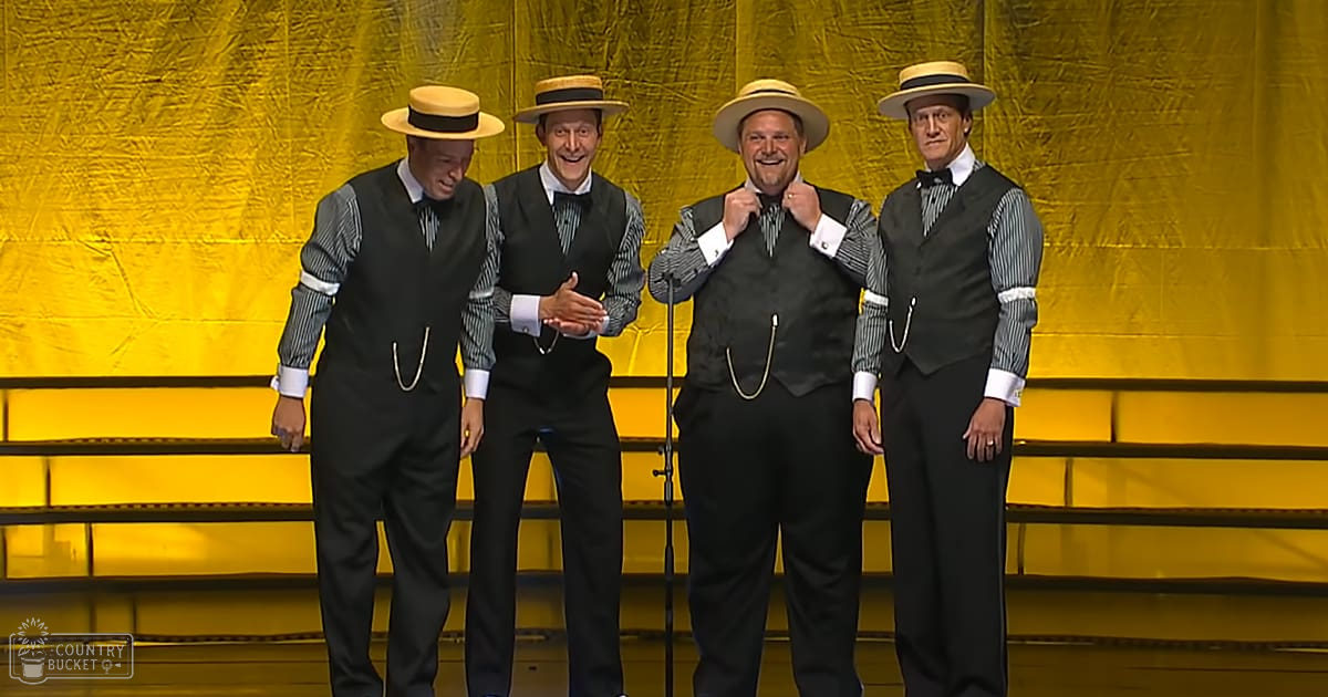Group performance scene at awards show or similar event; adult males in striped shirts, black vests, dark trousers, straw hats, bow ties; bright yellow backdrop, microphone stand, stage lighting suggests evening or night setting.