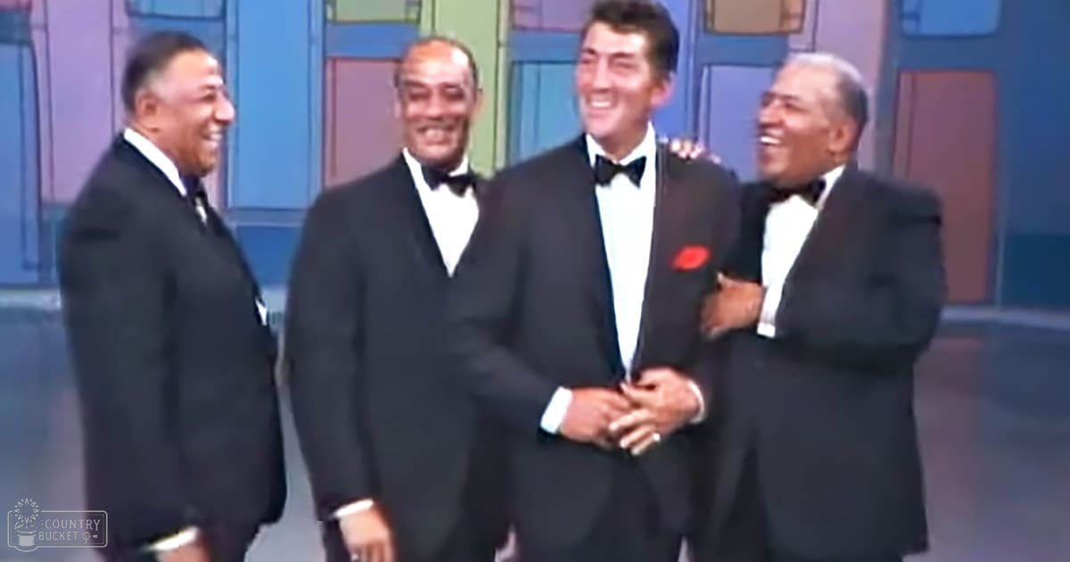 A group of adult male singers in formal attire from a past era, possibly performing on stage; setting is an indoor studio or theater with painted backdrop and artificial lighting.