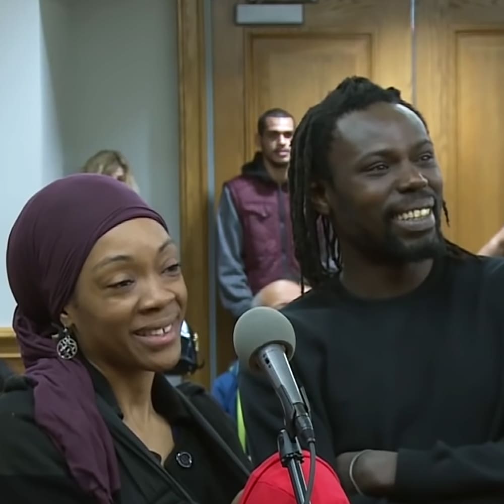 A young adult male with dreadlocks smiles at someone off-camera, surrounded by individuals and a wooden indoor setting.