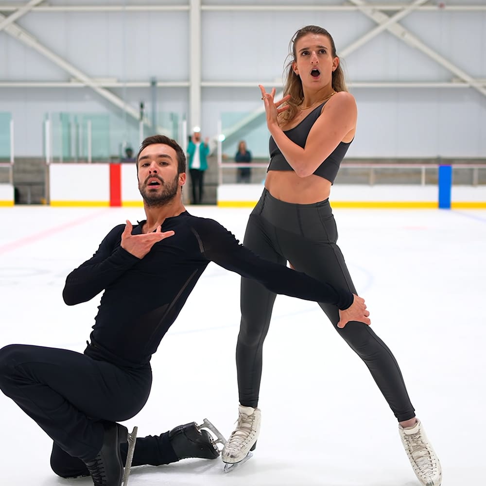 Young adult figure skating pair practicing at a well-lit indoor ice rink with spectators in the background.