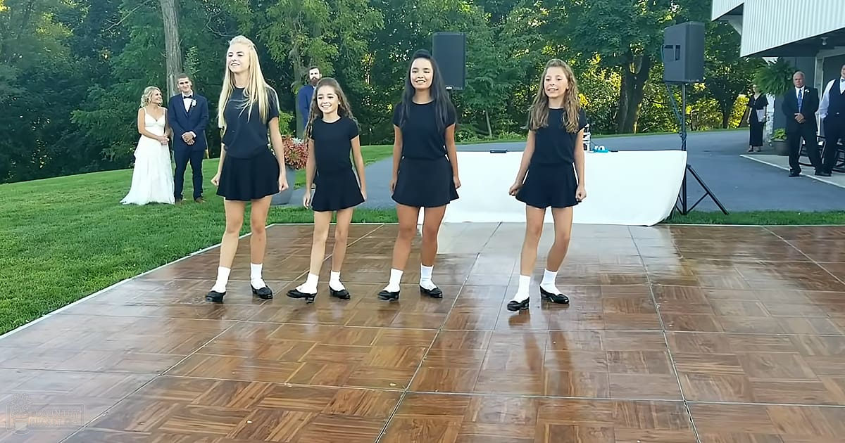 A group of young dancers in black dresses perform outdoors at an event with a bride and groom in formal attire standing in the background, under natural daylight.