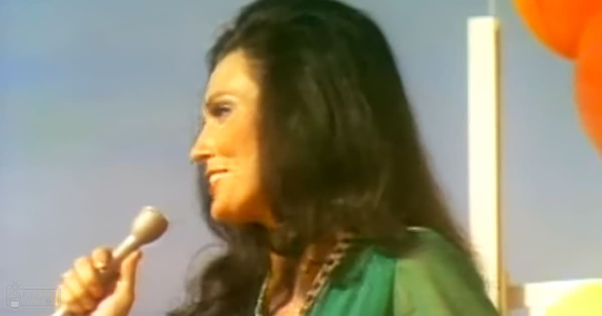 A woman speaking into a microphone at what appears to be an indoor event, wearing a green dress or top with long sleeves and a V-neckline. The setting is simple with a plain wall in the background.