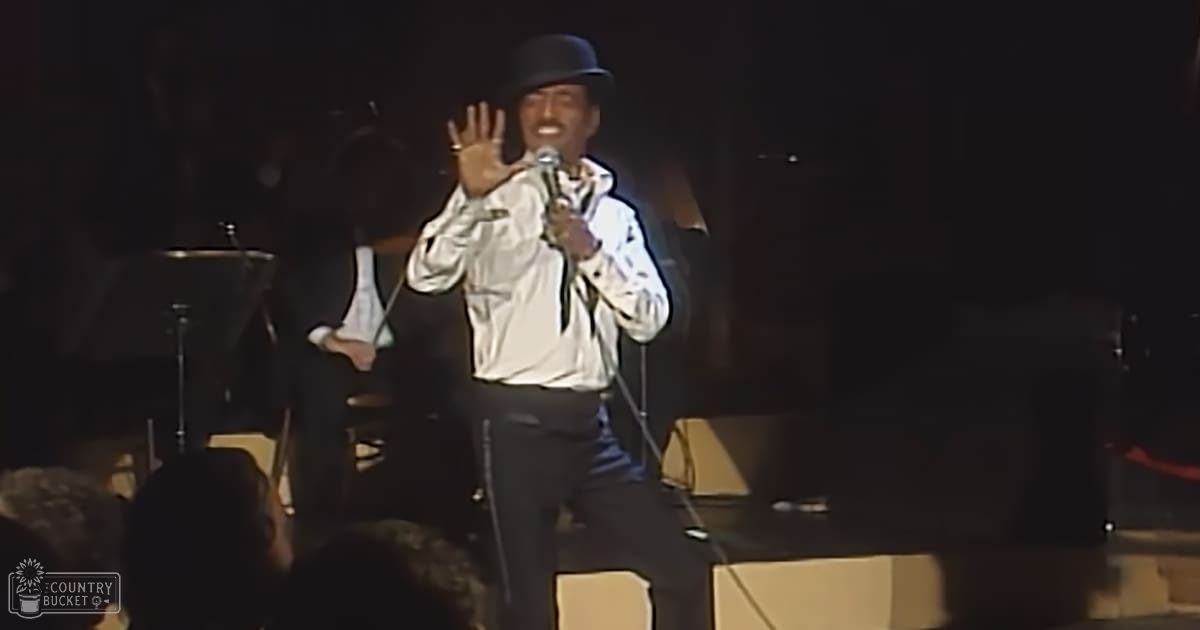 An adult male singer wearing a white jacket with black accents and a bowler hat, holding a microphone in an indoor concert setting.