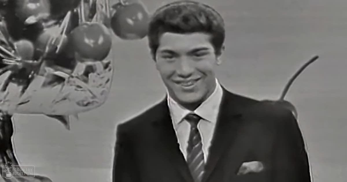 A young adult male wearing a dark suit jacket and white dress shirt with a tie stands indoors against a neutral backdrop.
