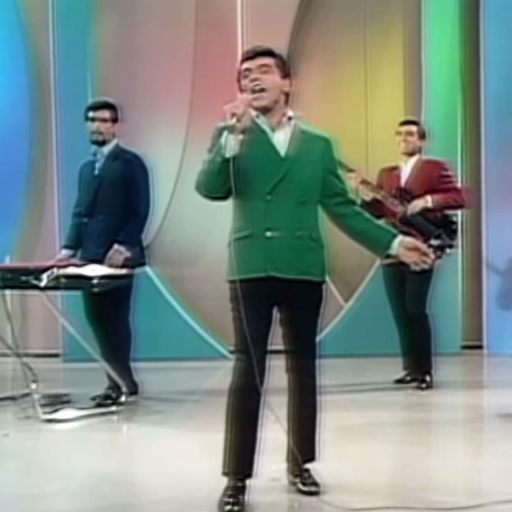An adult male performer in a green double-breasted suit with two backup musicians in dark mid-20th century-style suits on stage, set against a colorful abstract backdrop.