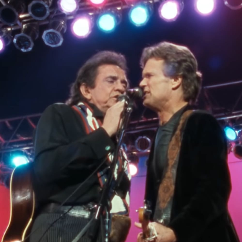 Middle-aged male performers in a concert setting, wearing patterned jackets with microphones and guitars.