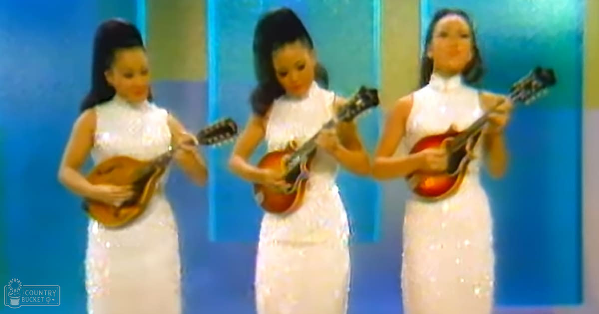 Three young adult female musicians perform with mandolins against a blue backdrop on an indoor stage.