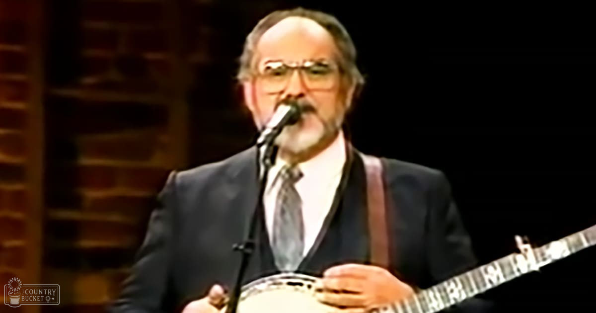 A middle-aged male musician in a dark suit plays a banjo in front of a microphone on stage at an evening concert.