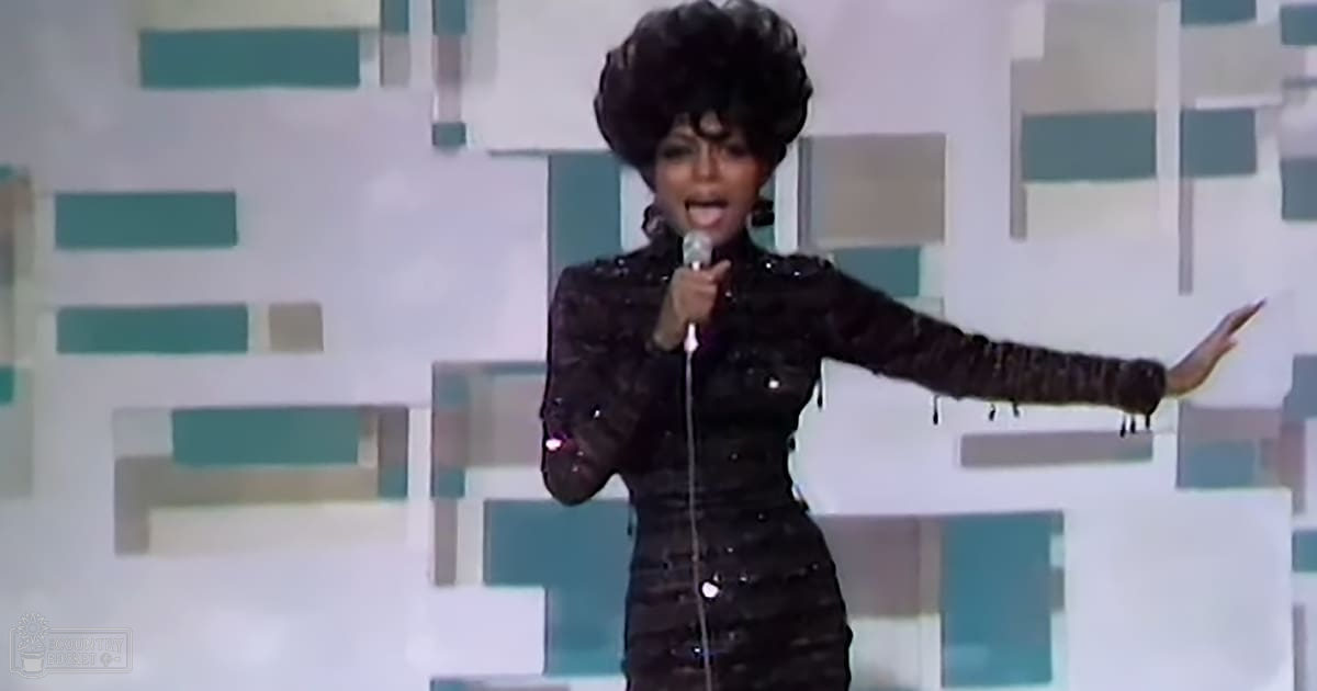 A female performer wearing a black, shiny dress with embellishments and a high neckline stands on an indoor stage with a modernistic geometric backdrop. The setting suggests a daytime performance or recording session.