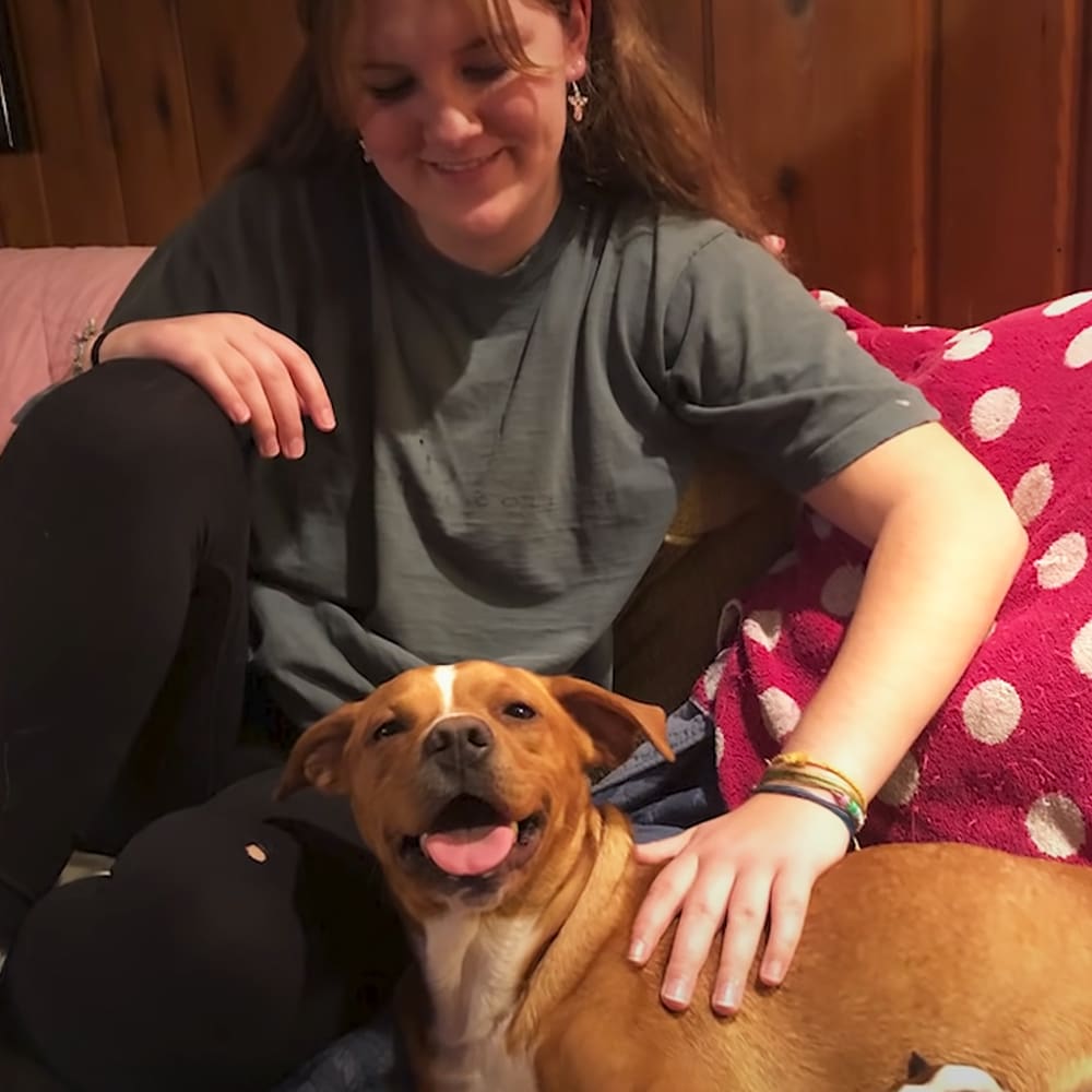 A young adult female wearing a gray t-shirt, dark pants, and colorful bracelets sits with her brown and white mixed breed dog in a cozy indoor setting. A red polka-dotted blanket is visible on the furniture they are sitting on.