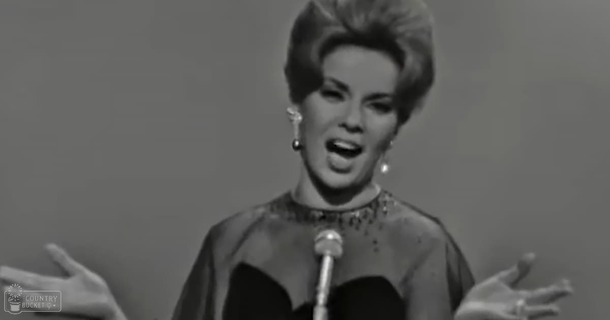 An adult female singer wearing an elegant vintage-style dress with decorative elements, holding a microphone in a studio setting.