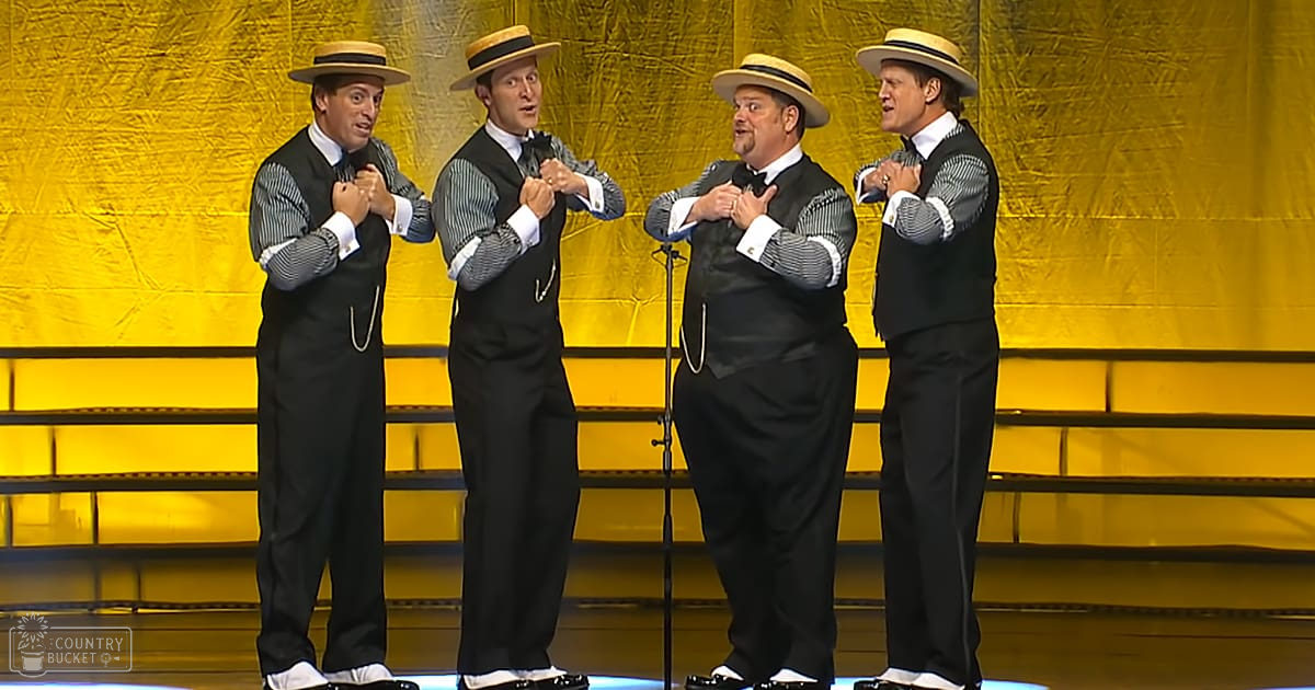 Four middle-aged male performers wearing striped shirts, black vests, white collared shirts underneath, black trousers, white shoes, and straw hats on a stage with bright yellow curtains.