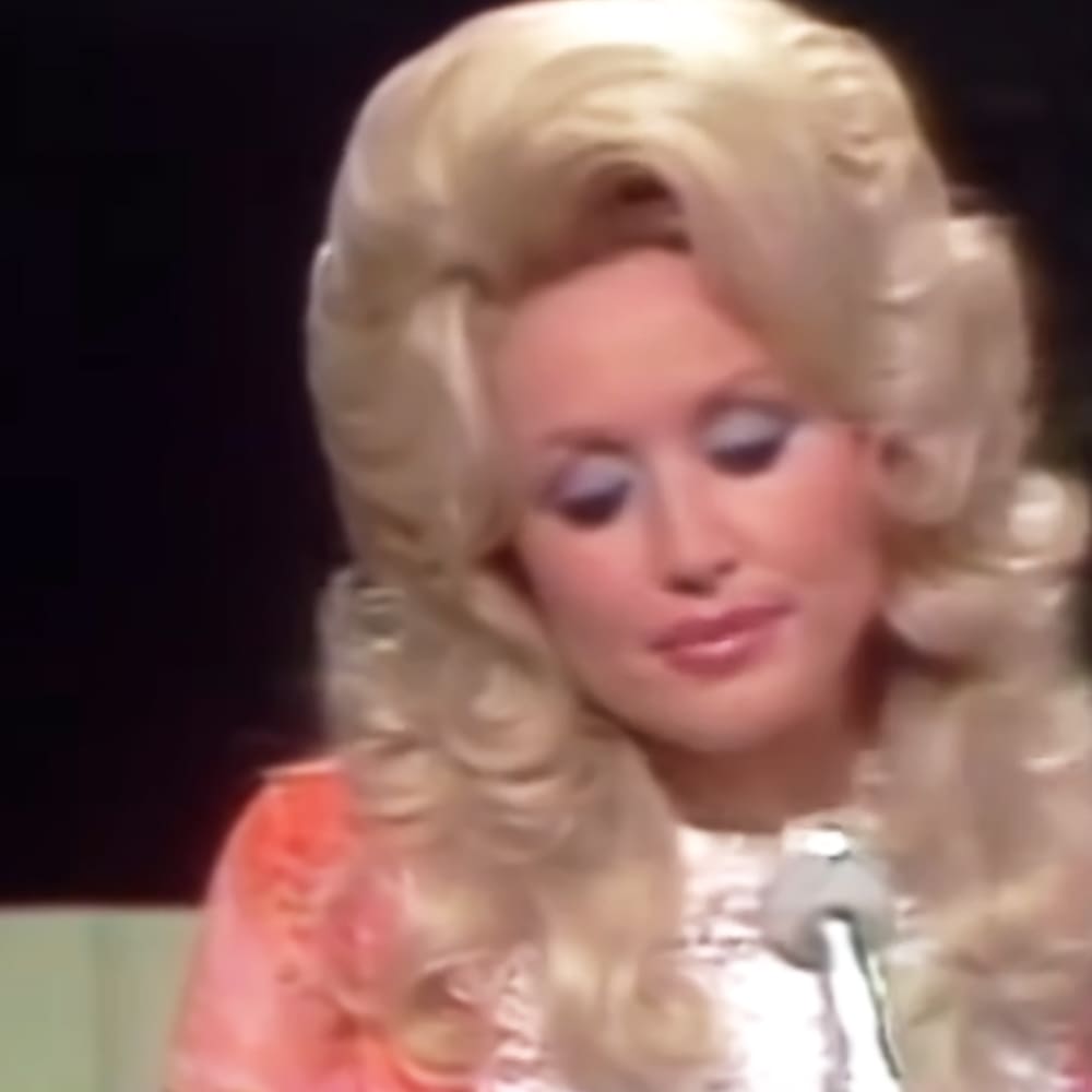 A woman with light blonde hair, wearing a white underlayer and a pinkish-orange floral ruffled garment from the 1970s or early 1980s style, stands in a dimly lit indoor setting.