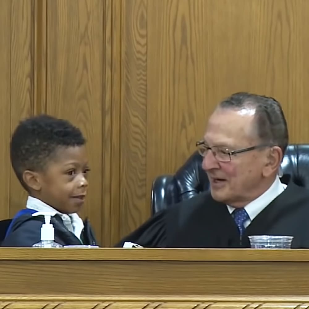 A toddler in a courtroom, wearing a dark jacket with white trim and a blue collar, seated beside an older adult male in formal judicial attire.