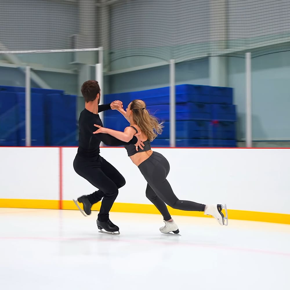 A young couple skating together in an indoor ice rink with blue padded walls and glass barriers, under even lighting suggesting daytime or early evening.