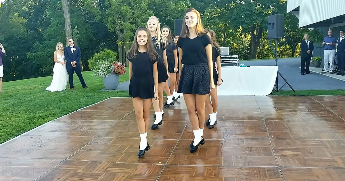 A group of young dancers performing outdoors at what seems to be a wedding reception with a bride and groom in the background.