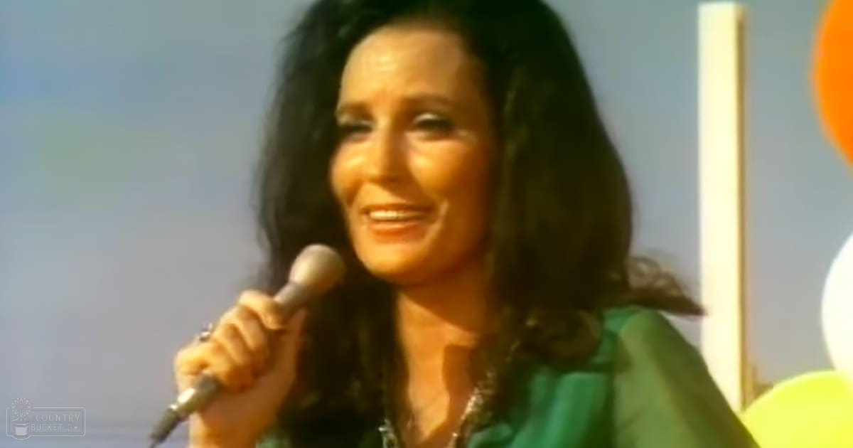 A middle-aged female wearing a green 1970s-style V-neck garment indoors, holding a microphone with yellow and orange balloons in the background.
