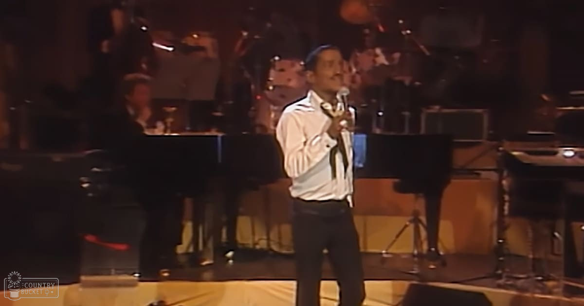 An adult male singer performing on stage under dim lighting with a piano and drums visible in the background of an indoor concert setting.