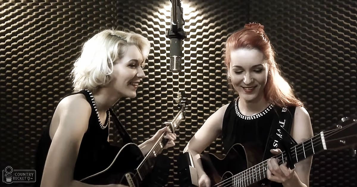 Two adult female musicians with pearl necklaces and form-fitting black tops, holding guitars, in a recording studio setting with acoustic foam panels and professional lighting.
