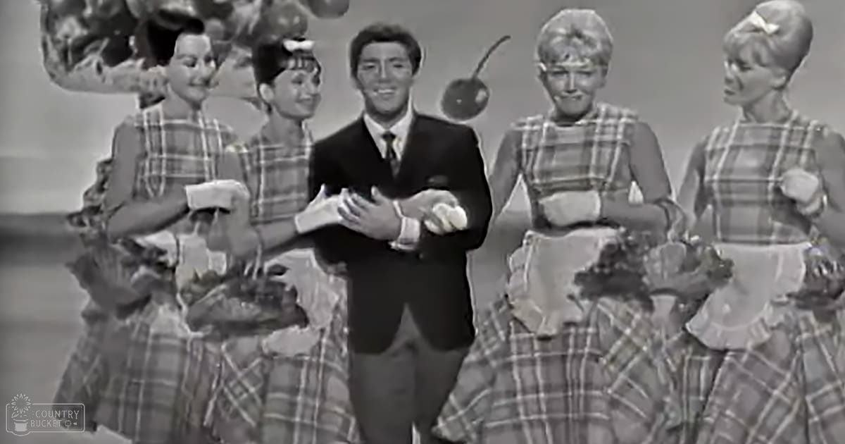 A man in a formal dark suit jacket and white shirt performing with a group of young women dressed in checkered dresses in a studio setting.