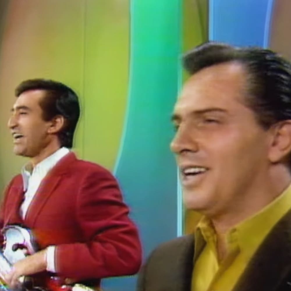Two adult males in a studio setting with colorful panels behind them, wearing blazers from the mid-20th century.
