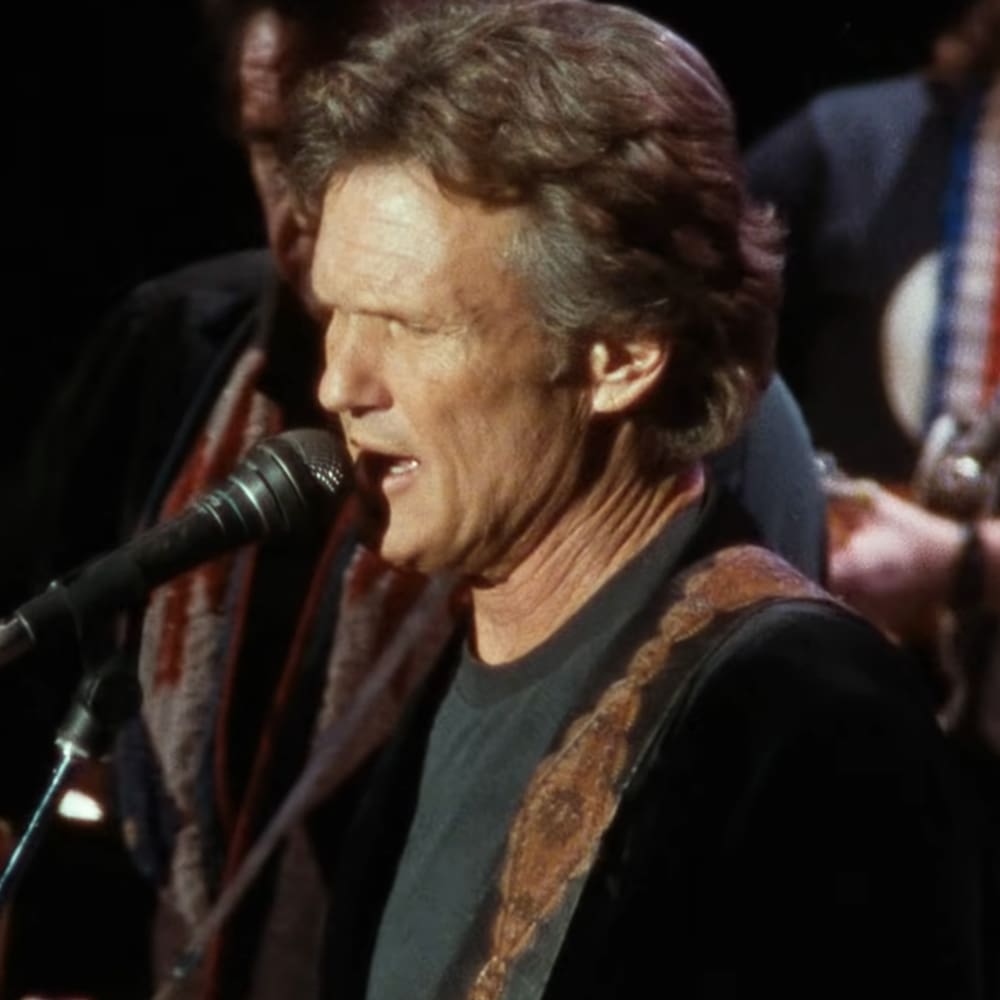Middle-aged man in a black t-shirt and dark-colored jacket with a patterned strap, performing at an indoor concert or event; another adult male wearing a red and white striped shirt and lanyard stands nearby.