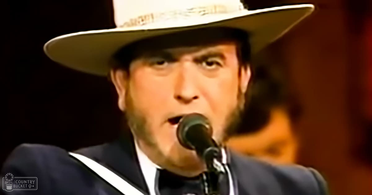 An adult male performer in formal mid-20th-century attire with a white cowboy hat and dark suit jacket, holding a microphone on stage under focused lighting.