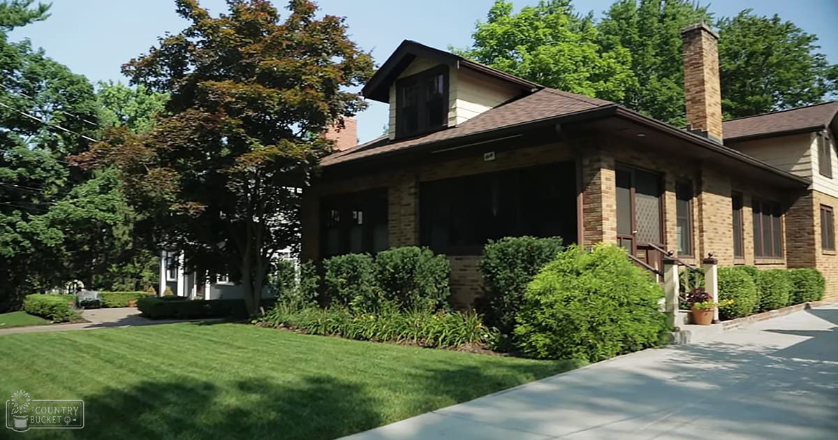 A well-maintained suburban landscape with a brick and siding house, a lawn bordered by hedges and trees, and mature shade-providing trees in spring or summer.