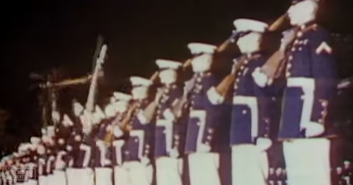 A nighttime military parade with young adult males in dark blue uniforms, white pants, and caps, holding bugles under artificial lighting against a dark background.