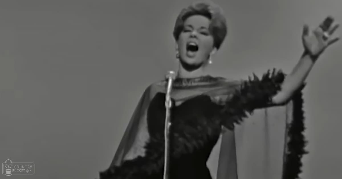 A female adult singer in a black, feather-adorned dress on stage with a microphone stand and light-colored background.