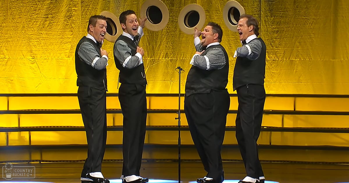 A group of adult male singers performing on a stage with yellow backdrop and circular cutouts, possibly during an indoor concert or theater performance at evening lighting.
