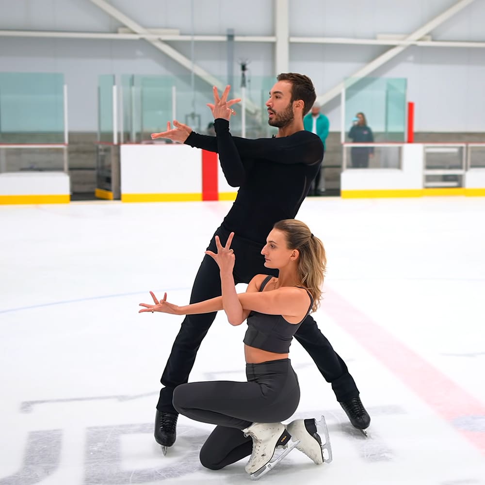Young adult male figure skater in a black long-sleeve leotard and tight-fitting pants practices with a female partner at an indoor ice rink under artificial lighting, surrounded by spectators.