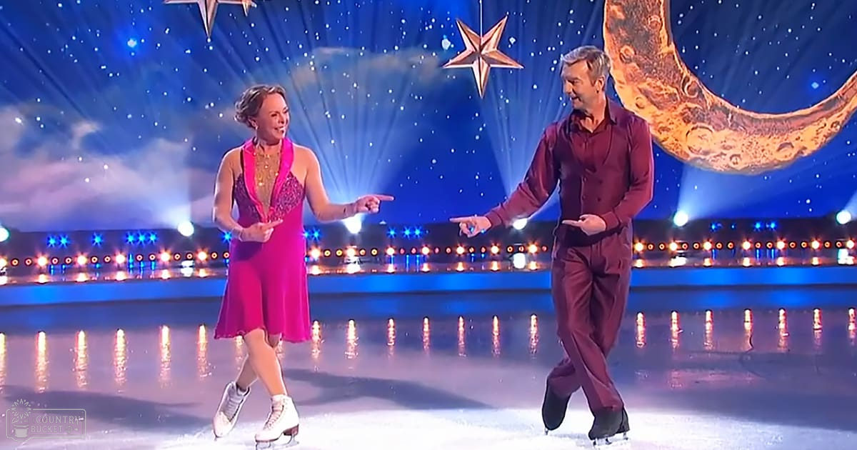 A female figure skater in a red, vintage-style dress with white figure skates performs alongside an adult male figure skater wearing a maroon shirt and trousers, on an indoor ice rink decorated with stars and a crescent moon under bright stage lights.