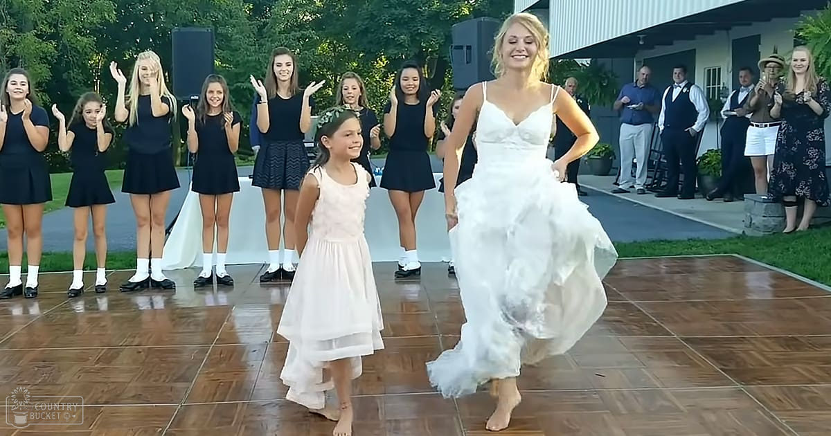 An outdoor wedding reception with a bride dancing with a young girl amidst greenery and wooden decking, under natural sunlight.