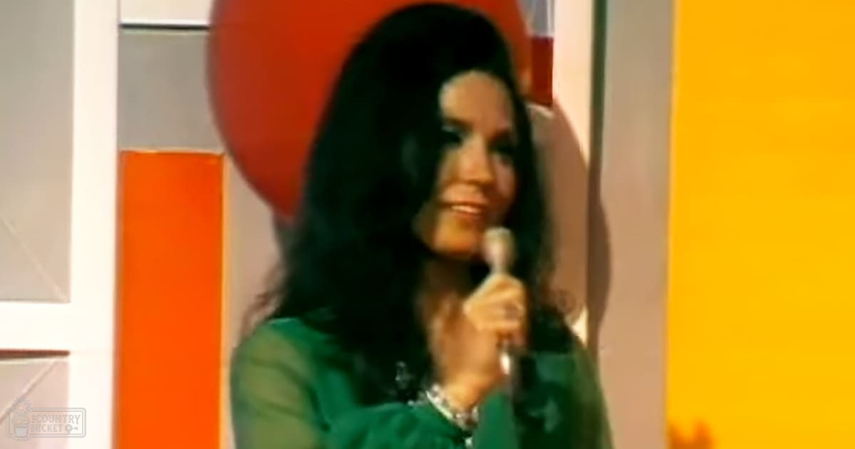 A middle-aged female wearing a green, smooth-silk or satin 1960s-era garment stands in an indoor setting with a microphone and bright orange and yellow walls.
