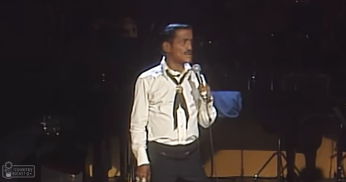 A middle-aged male performer wearing a white shirt with black accents and an undone tie, holding a microphone on stage at an indoor venue.