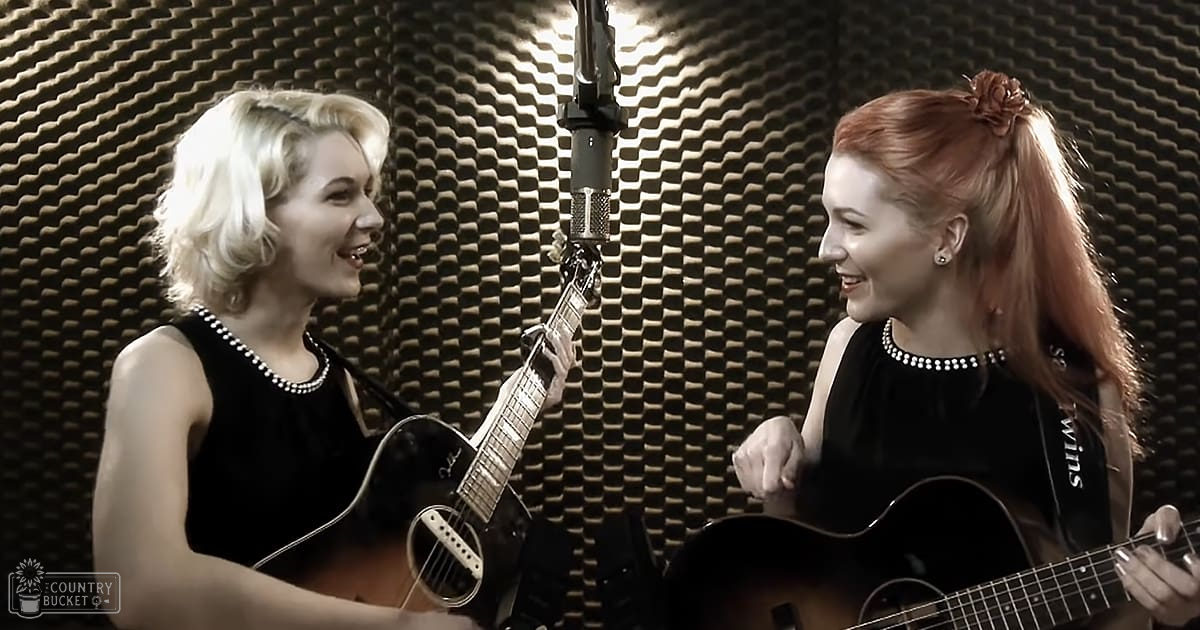 Two adult female singers in a recording studio; one with short blonde hair wearing a vintage black dress, another with long red hair adorned with a flower accessory and modern black attire. Microphone stand central, acoustic foam panels indicating soundproofed environment.