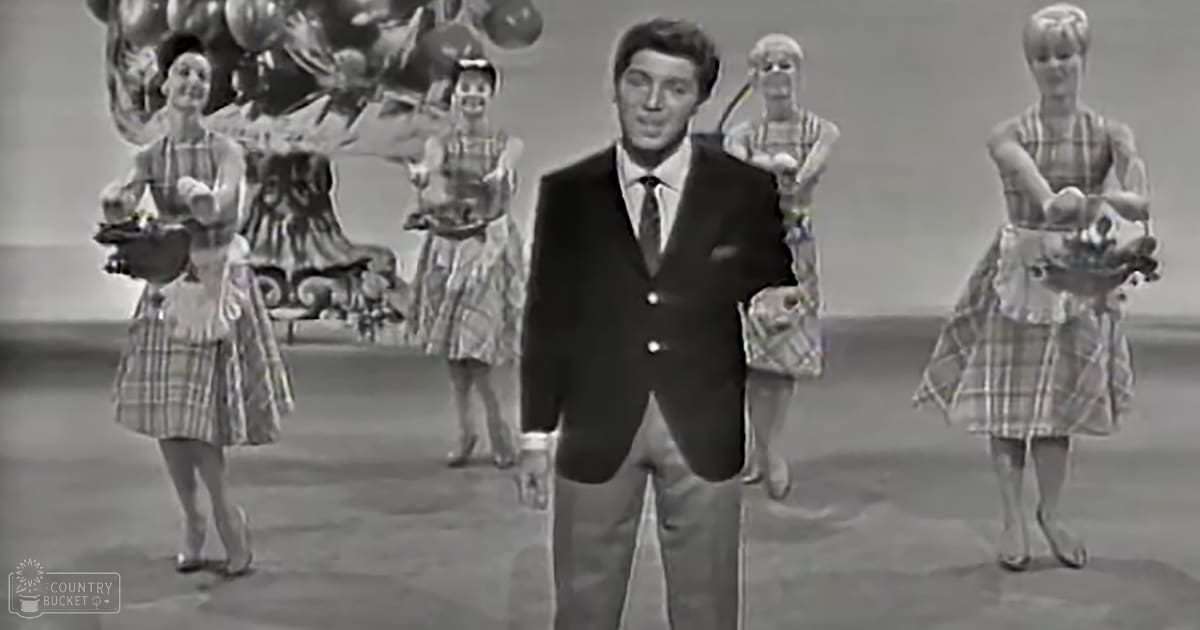 A middle-aged man dressed formally in a dark suit with a white shirt and tie stands on a stage performance set with decorative elements like a chandelier and balloons, suggesting an indoor festive occasion.