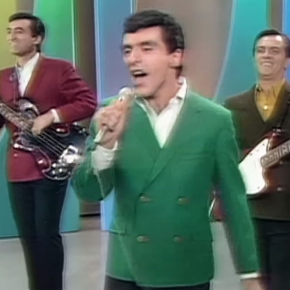 An adult male singer in a green double-breasted blazer, performing at an indoor stage with colorful abstract background panels and holding a guitar.